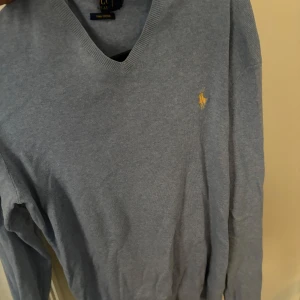 Blå v-ringad tröja från Ralph Lauren - Säljer en stilren blå v-ringad tröja från Ralph Lauren med det klassiska logotypbroderiet på bröstet. Tröjan har långa ärmar och är perfekt för både vardag och mer uppklädda tillfällen.