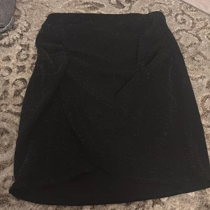 Svart glittrig kjol från Kappahl - Snygg svart kjol från Kappahl med glittrande detaljer. Perfekt för att lysa upp din outfit. Kjolen har en enkel och stilren design som passar till många olika tillfällen.