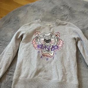Grå sweatshirt från Kenzo med tigertryck - Stl 14 år. Liten i storlek passar upp till 10 år. Säljer en grå sweatshirt från Kenzo med ett coolt tigertryck i flera färger på framsidan. Tröjan har långa ärmar och ribbade muddar vid ärmslut och nederkant. Perfekt för en avslappnad stil.