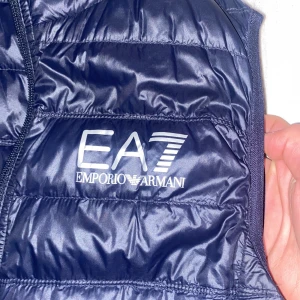 Mörkblå väst från Emporio Armani - Snygg mörkblå dunväst från Emporio Armani EA7 med dragkedja och quiltad design. Köpt från boozt, storlek. Är som nyskick men bara att lappen inne i västen är skrynklig. 