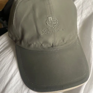 Grön keps från Loro Piana - Snygg grön keps från Loro Piana med broderad logotyp framtill. Kepsen har en klassisk design och är tillverkad med Storm System-teknologi för extra skydd. Insidan har ett stilrent rutigt mönster.