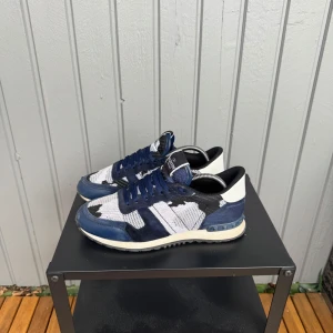 Valentino sneakers i blått och vitt - Snygga Valentino sneakers i blått och vitt med ett unikt camouflage-mönster. Skorna har snörning och en bekväm sula, perfekt för en stilren look. De är tillverkade i en kombination av material som ger både stil och komfort.