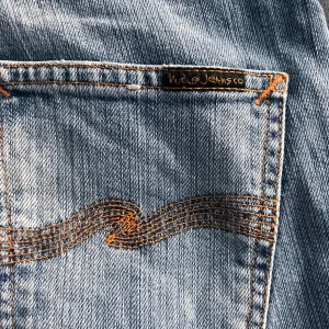 Blå jeans från Nudie Jeans - Säljer ett par snygga blå jeans från Nudie Jeans med klassisk femficksdesign och orange sömmar. Jeansen har en broderad detalj på bakfickan och är tillverkade av ekologisk bomull. Perfekta för dig som gillar en stilren look med en twist.