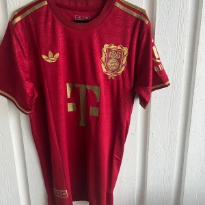 Röd fotbollströja från Adidas - Säljer en röd fotbollströja från Adidas med guldiga detaljer och tryck. Tröjan har korta ärmar och ett snyggt emblem på bröstet. Perfekt för fotbollsfans som vill visa sitt stöd.