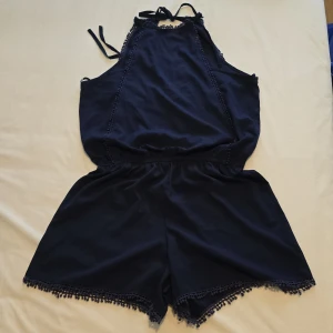 Mörkblå jumpsuit med spetsdetaljer - Snygg mörkblå jumpsuit med spetsdetaljer längs kanterna. Den har en v-ringad hals och knytband vid axlarna. Perfekt för en somrig look!