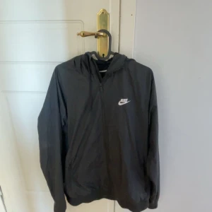 Svart vindjacka från Nike - Säljer en stilren svart vindjacka från Nike med dragkedja och huva. Jackan har en klassisk design med Nike-logga på bröstet och är perfekt för blåsiga dagar. Den är långärmad och har en bekväm passform. Passar perfekt nu inför golf starten.