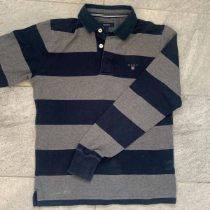 Randig pikétröja från GANT - Säljer en stilren randig pikétröja från GANT i mörkblått och grått. Tröjan har långa ärmar och klassisk krage med knappar. Perfekt för en avslappnad men ändå snygg look.