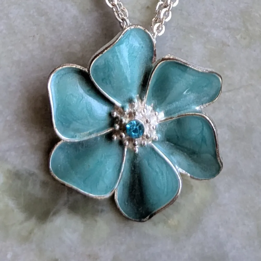 Charmigt halsband med en stor blomma. Blomman har en glittrande blå sten i mitten och hänger på en elegant kedja. Perfekt för att ge en färgklick till din outfit.. Asusteet.