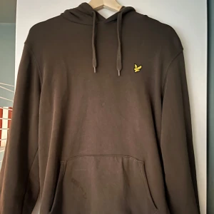 Oliv färgad hoodie från Lyle & Scott - Säljer en Oliv färgad hoodie från Lyle & Scott med en gul logga på bröstet. Tröjan har en klassisk känguruficka och justerbar huva med dragsko. Perfekt för en avslappnad stil.