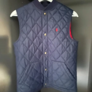 Blå quiltad väst från Polo Ralph Lauren - Snygg blå quiltad väst från Polo Ralph Lauren med knappar framtill. En liten logga på bröstet. Perfekt för lager-på-lager-stil. Västen passar prima till dig som är runt 170-175..