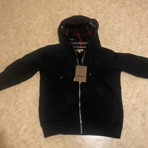 Svart huvtröja med dragkedja från Burberry - Snygg svart huvtröja från Burberry med dragkedja och klassiskt rutigt mönster på insidan av huvan. Perfekt för en stilren look med en touch av lyx. Huvan har snörning för justering.armand får ni med om ni köper cordell rostfritt stål