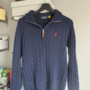 Blå stickad tröja från Polo Ralph Lauren - Snygg blå stickad tröja från Polo Ralph Lauren med dragkedja och kabelstickat mönster. Tröjan har en klassisk krage och den röda logan på bröstet. Perfekt för kyligare dagar. Den är tidigare köpt från Vinted men skicket känns i princip helt nytt.