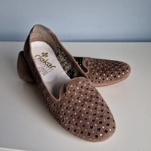 Bruna loafers från Rieker - Snygga bruna ballerina/loafers från Rieker i mocka med ett perforerat mönster och strass. Skorna har en bekväm passform och är perfekta för en stilren look.