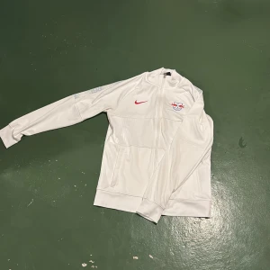 Vit träningsjacka från Nike Rb Leipzig  - Säljer en stilren vit träningsjacka från Nike med dragkedja och RB Leipzig-logga på bröstet. Jackan har långa ärmar och en röd Nike-logga. Perfekt för sportiga aktiviteter eller som en snygg vardagsjacka.