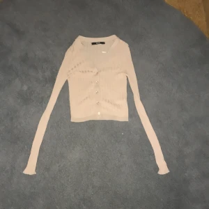 Beige/rosa ribbad kofta med knappar - Säljer en stilren beige kofta med ribbad struktur och knappar framtill. Perfekt för lager-på-lager och passar till många olika outfits. Långärmad design som ger en elegant look.