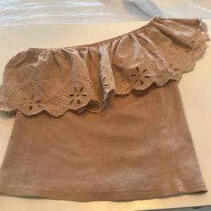 Beige offshoulder topp med spetsdetaljer - Säljer en söt beige offshoulder topp med vacker spetsdetalj längs kanten. Perfekt för varma sommardagar eller en avslappnad kväll. Toppen har en lös passform och är tillverkad i ett mjukt material.