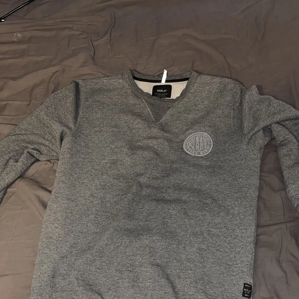 Säljer en stilren grå tröja från Replay med rund hals och långärmade ärmar. Tröjan har en broderad logotyp på bröstet och en liten etikett vid nederkanten. Perfekt för en avslappnad look.. Neuletakit & Villapaidat.