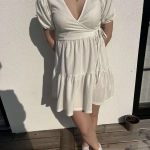 Vit omlottklänning från Gina Tricot - Säljer en söt vit omlottklänning med korta ärmar och knytning i midjan. Klänningen har en v-ringad design och volanger som ger en romantisk känsla. Perfekt för varma sommardagar!