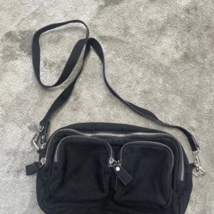 Svart axelväska med dragkedjor - Säljer en svart axelväska med flera dragkedjor och en justerbar axelrem. Väskan har två framfickor med dragkedja och en dekorativ rem med nitar.