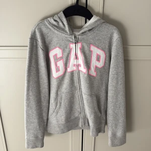 Grå hoodie från GAP Kids - Säljer en grå hoodie från GAP Kids med rosa detaljer. Den har en dragkedja framtill och en stor logga på bröstet. Perfekt för en avslappnad stil. 🩶