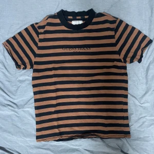 Randig t-shirt från Guess Jeans storlek Medium - Säljer en cool randig t-shirt från Guess Jeans i svart och brun. Den har korta ärmar och en klassisk rund halsringning. Perfekt för en avslappnad stil. Storlek Medium passar också small tycker jag