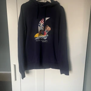 Mörkblå hoodie från Polo Ralph Lauren - Säljer en snygg mörkblå hoodie från Polo Ralph Lauren med ett tryck av en björn som seglar. Perfekt för en avslappnad stil. Hoodien har långa ärmar och en klassisk huva.
