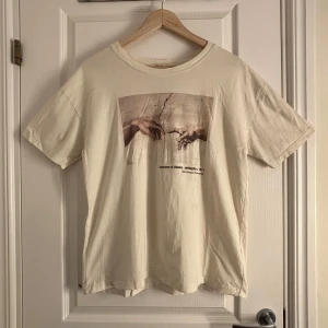 Beige t-shirt med konsttryck från Pull&Bear - Säljer en beige t-shirt från Pull&Bear med ett konsttryck inspirerat av Michelangelos 'Skapelsen av Adam'. T-shirten har en normal passform och är kortärmad, perfekt för en avslappnad stil. Tillverkad i bomull för en skön känsla. Passar bra till jeans eller shorts!