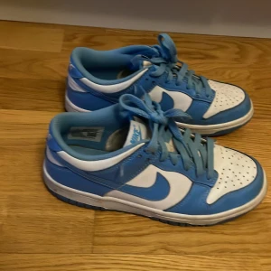 Nike Dunk Low blå/vita   - Nike Dunk Low sneakers i blått och vitt skinn med klassisk swoosh på sidorna. Lågt skaft, perforerad tå och snörning framtill. Snygg kontrast mellan de blå och vita panelerna, perfekt för dig som gillar streetstyle och vill sticka ut.
