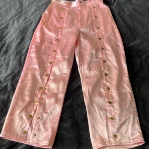 Rosa satinbyxor från Nikolaj Storm - Glansiga rosa byxor från Nikolaj Storm i satinliknande material. Byxorna har raka ben, hög midja och coola tryckknappar längs sidorna. Två bakfickor och hällor för bälte. Perfekta för dig som vill sticka ut med en unik vibe.