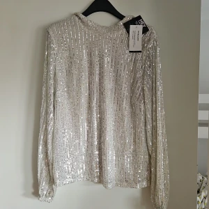 Glittrig silverblus. - Superfin glittrig blus från nelly i silver med långa ärmar och paljetter över hela. Blusen har en rak passform och hög krage, perfekt för att sticka ut. Fodrad på insidan för extra komfort. Aldrig använd. 