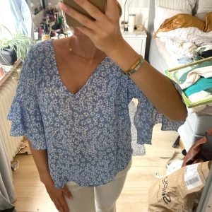 Blommig blå blus med volangärm - Supersöt blommig blus i ljusblått med vita blommor. V-ringad och med vida volangärmar som ger en luftig känsla. Perfekt för dig som gillar romantiska och somriga vibes. Tunn och skön, passar perfekt till jeans eller kjol.