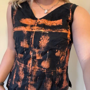 Svart och orange linnetopp med V-ringning - Unik svart linnetopp med coolt orange abstrakt mönster framtill. Toppen har V-ringning både fram och bak och är ärmlös, vilket ger en edgy och modern vibe. Perfekt för dig som vill sticka ut med en kreativ och konstnärlig look.