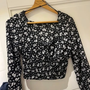 Svart blommig blus från BikBok  - Supersöt svart blus från BikBok  med vitt blommönster. Modellen är croppad med smockad midja, v-ringning och långa ärmar. Stl xs