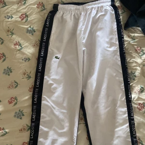Vita Lacoste träningsbyxor med logga - Säljer ett par vita träningsbyxor från Lacoste i storlek S. Byxorna har svarta sidor med Lacoste-tryck längs benen och klassisk broderad krokodil på framsidan. Resår i midjan och sportig look, perfekta för chill eller träning.