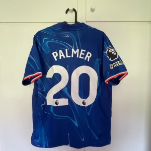 Chelsea Palmer 20 matchtröja Nike - Chelsea FC hemmatröja med Palmer 20-tryck. Blå bas med unikt mönster i ljusare blå toner, röda och vita detaljer på ärmsluten. Broderad Chelsea-logga och Nike swoosh framtill, Premier League-märke på ärmen. Tillverkad i lätt och ventilerande polyester.