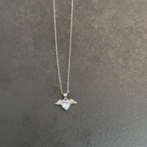 Halsband med hjärta och vingar - Snyggt halsband i silverfärg med ett gnistrande hjärta i mitten och små vingar på sidorna. Perfekt accessoar för dig som gillar unika detaljer och vill sticka ut lite extra.