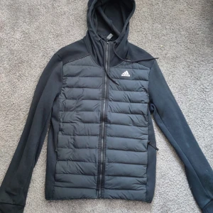 Svart pufferjacka med huva från Adidas - Svart pufferjacka från Adidas med quiltad front, huva och dragkedja hela vägen. Jackan har långa ärmar i mjukt material och två sidofickor med dragkedja. Perfekt för dig som gillar sportig stil och vill ha något som funkar till vardags.