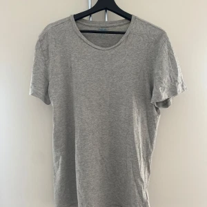 Grå Ralph Lauren T-shirt - En stilren grå t-shirt från Polo Ralph Lauren med klassisk rund hals och korta ärmar. T-shirten har en liten broderad logga i mörkblått nedtill. Tillverkad i mjuk bomull för en skön känsla och enkel att matcha med allt.