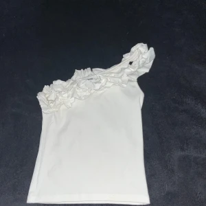 Vit oneshoulder topp med volang Kappahl - Supersöt vit oneshoulder topp från Kappahl med stora volangdetaljer längs axeln. Topp i mjuk bomullskänsla, perfekt för dig som vill sticka ut lite extra. Enkel att matcha med både jeans och kjol.