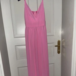 Lång rosa klänning med v-ringning - Säljer en lång, ljusrosa klänning med smala axelband och djup v-ringning framtill. Klänningen har ett markerat midjeband och ett luftigt, fallande tyg som ger en elegant siluett. Perfekt för sommarens speciella tillfällen.