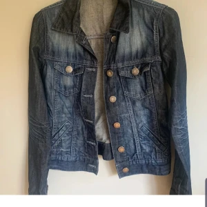 Klassisk blå jeansjacka med knappar - Snygg blå jeansjacka från Oasis med slitna detaljer och silverfärgade knappar. Jackan har två bröstfickor med lock och knapp, samt två sidofickor. Perfekt till lager-på-lager och funkar året runt.