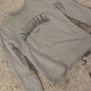 Grå sweatshirt med AMOUR-tryck - Snygg grå sweatshirt med texten AMOUR i glittriga bokstäver framtill. Tröjan har rund halsringning och långa ärmar, perfekt för en chill och avslappnad stil. Passar dig som gillar statement-plagg med lite extra bling.