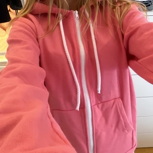 Rosa hoodie med dragkedja och huva - Säljer en rosa hoodie med vit dragkedja och snören i huvan. Tröjan har stora fickor framtill och är långärmad. Perfekt för dig som gillar en chill och avslappnad stil.