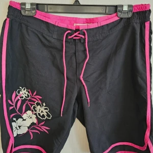 Svarta blommiga badshorts med rosa detaljer - Svarta badshorts med Hawaii tema med rosa kanter och snörning i midjan. Snyggt blommönster i vitt och rosa framtill och på bakfickan. Perfekta för stranden eller poolen, med en skön passform och coola detaljer.