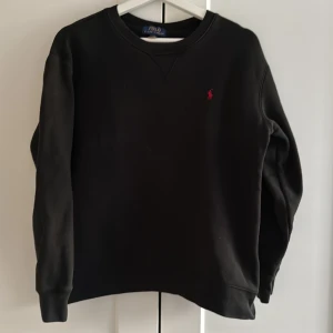 Polo Ralph Lauren Sweatshirt (Svart) - Svart sweatshirt från Polo Ralph Lauren med klassisk rund hals och broderad röd logga på bröstet. Tröjan har långa ärmar och ribbade muddar. Perfekt basic med clean look och skön passform.