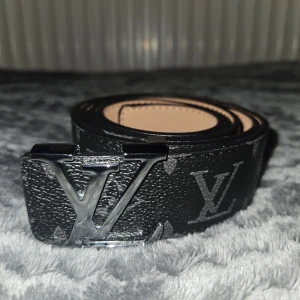Svart Louis Vuitton skärp i skinn - Snyggt svart skärp från Louis Vuitton med klassiskt LV-monogram och silverfärgat LV-spänne. Bältet är gjort i präglat skinn och har en stilren design med diskreta grå loggor längs hela bandet. Perfekt accessoar för att lyfta din outfit.