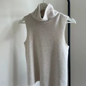 Beige polotopp från Zara - Säljer en stilren beige polotopp från Zara. Toppen är ärmlös med hög polokrage och har en mjuk, stickad känsla. Perfekt för lager-på-lager och enkel att matcha med olika outfits.