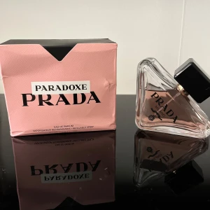 Prada Paradoxe Eau de Parfum - Prada Paradoxe Eau de Parfum med en unik trekantig glasflaska och svart lock. Parfymen kommer i en snygg rosa kartong med svart och vit text. Flaskan har en modern design och innehållet är ljusrosa. Perfekt för dig som gillar exklusiva dofter och stilren design.