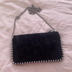 Svart axelväska med kedja och nitar - Snygg svart axelväska i mocka-look med silverfärgad kedjerem och coola nitar längs kanterna. Väskan har dragkedja upptill och flera fack inuti för att hålla ordning på dina grejer. Kedjan går att tas bort.