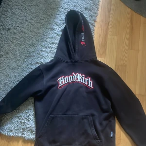 Svart Hoodrich hoodie med brodyr - Svart hoodie från Hoodrich med stor broderad logga på bröstet i vitt och rött. Texten 'From Nothing To Something' broderad på huvan. Klassisk känguruficka och lång ärm. Perfekt streetwear-vibe och riktigt snygga detaljer.